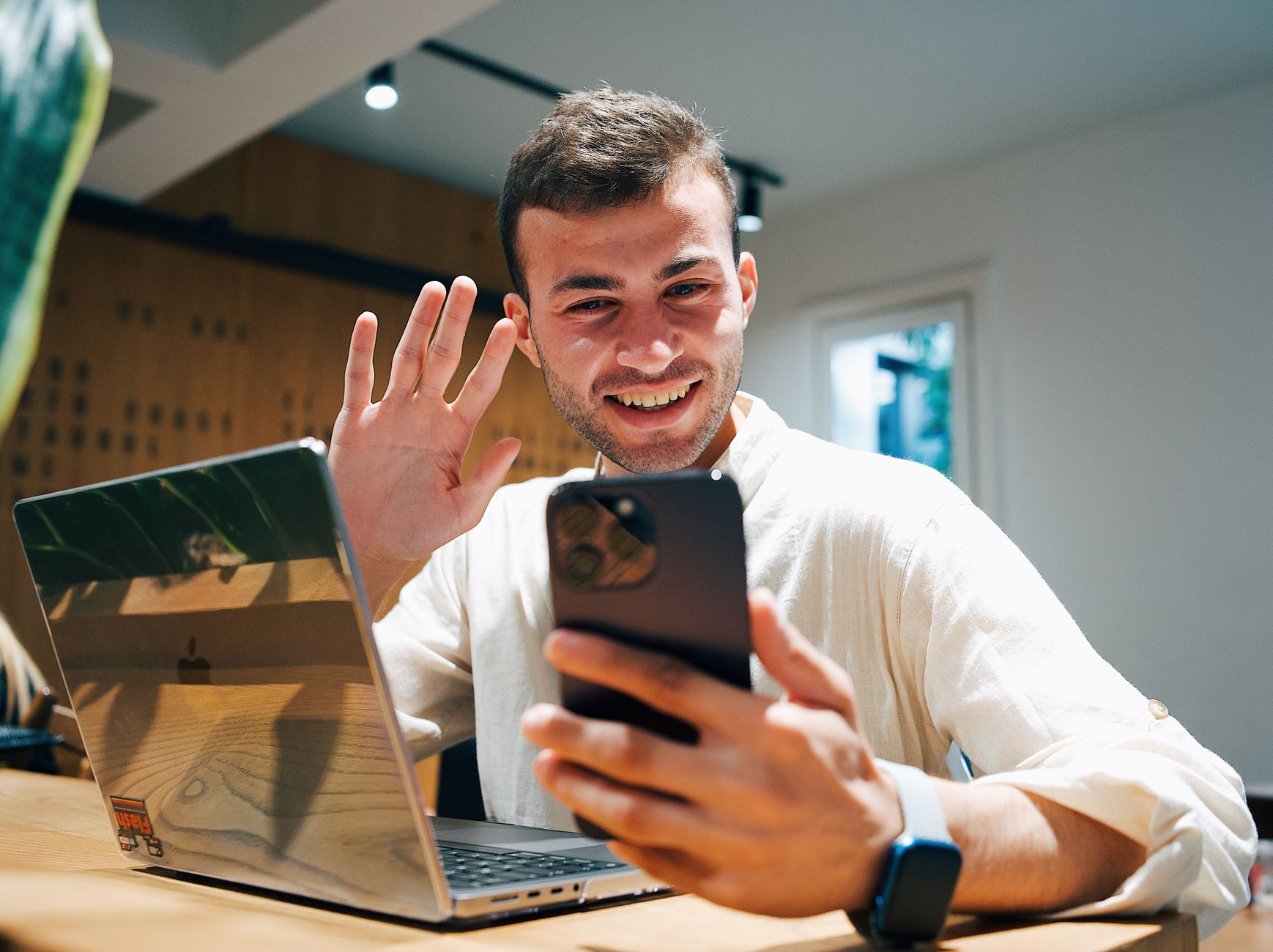 Hombre joven sonriente en videollamada con su smartphone junto a su laptop.